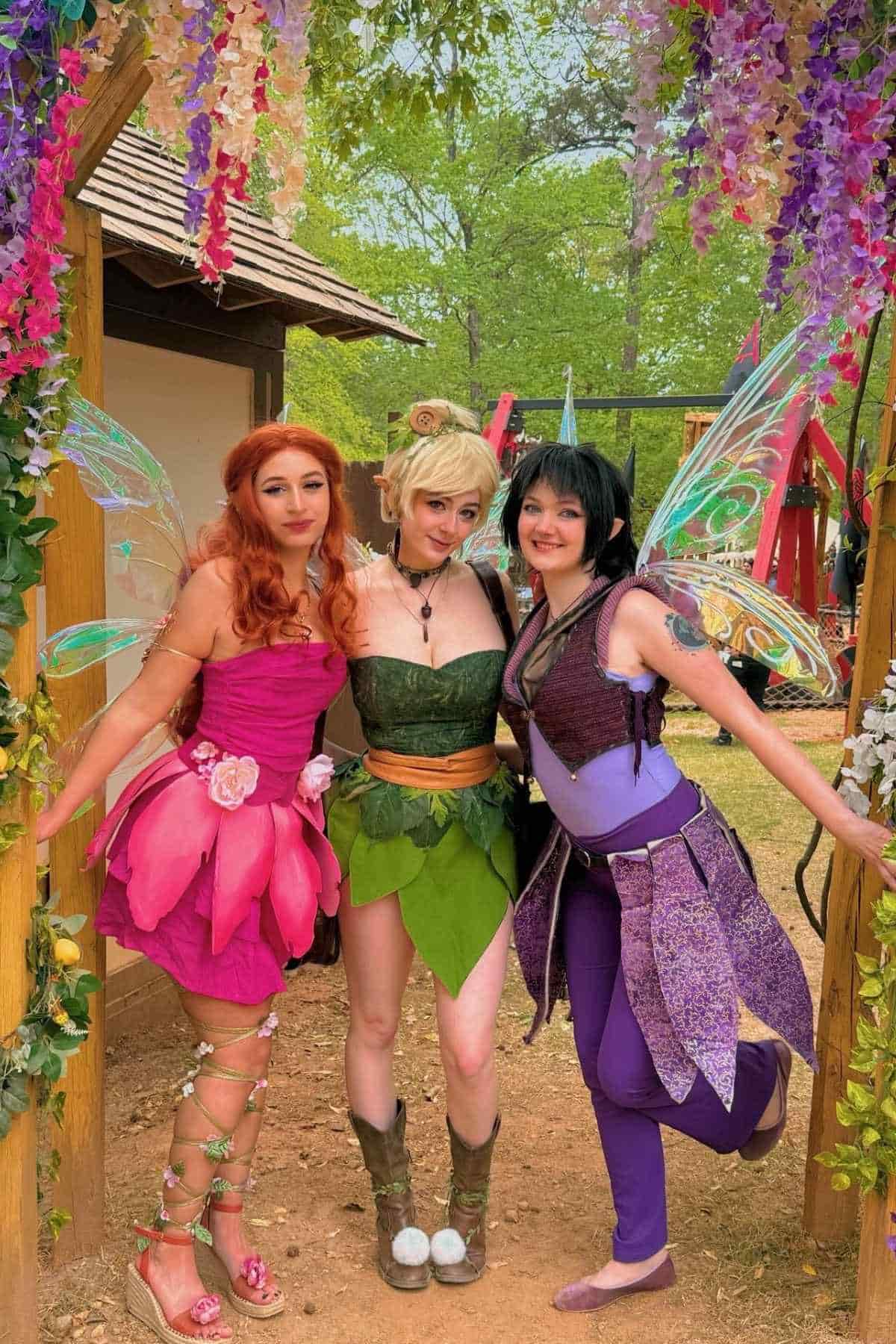 Three people in colorful fairy costumes pose together under a wooden arch decorated with flowers, with greenery in the background.