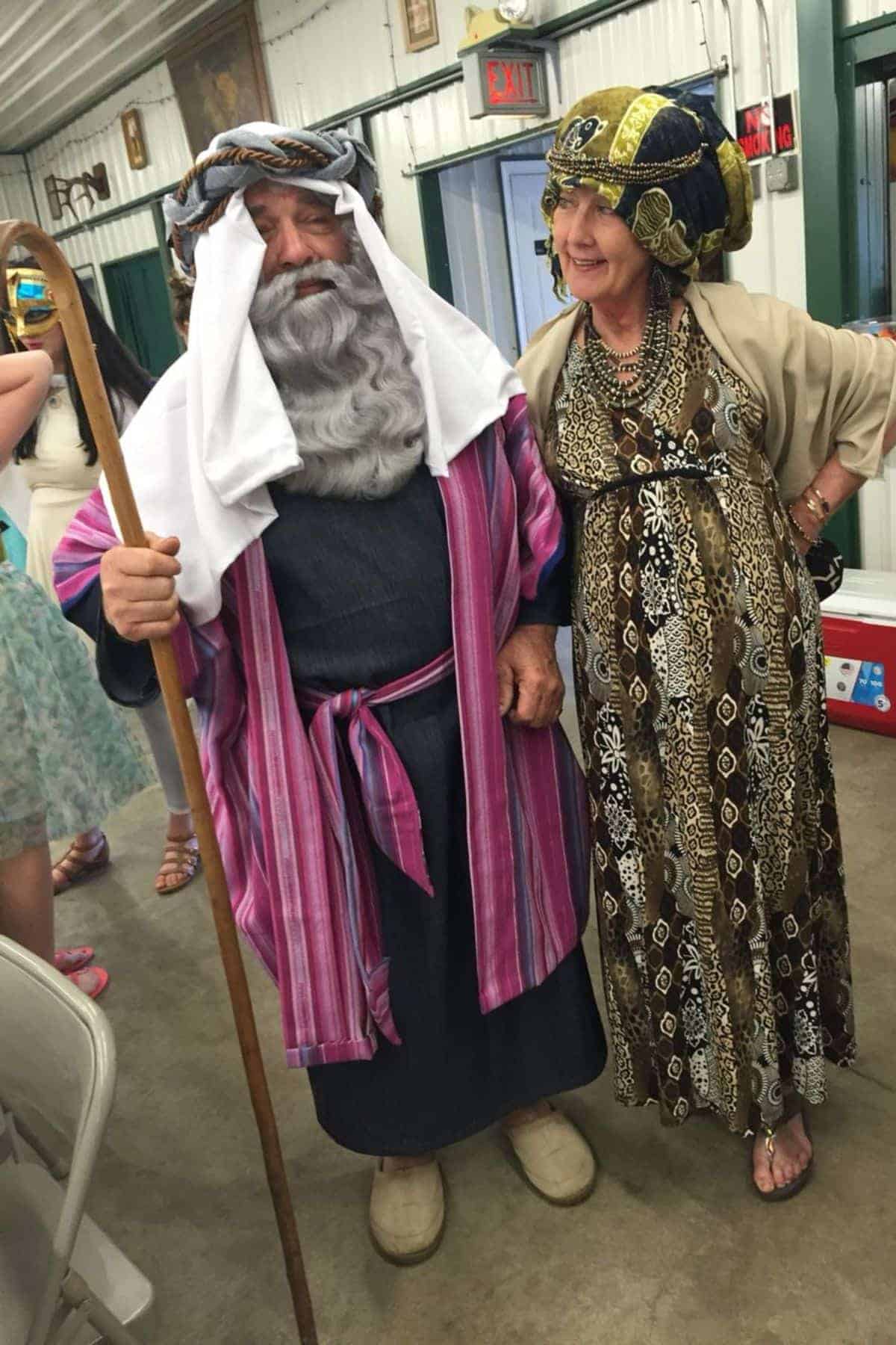 Two adults in biblical-era costumes stand indoors; the man holds a staff and wears a fake beard, while the woman wears a patterned dress and headpiece.