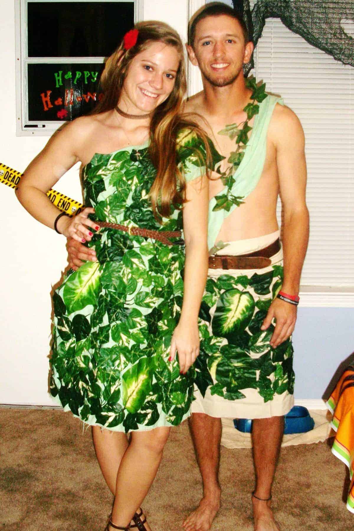 Two people wearing leaf-themed costumes stand together indoors. The woman has a flower in her hair; the man is shirtless with vines draped over his shoulder. Halloween decorations are visible behind them.