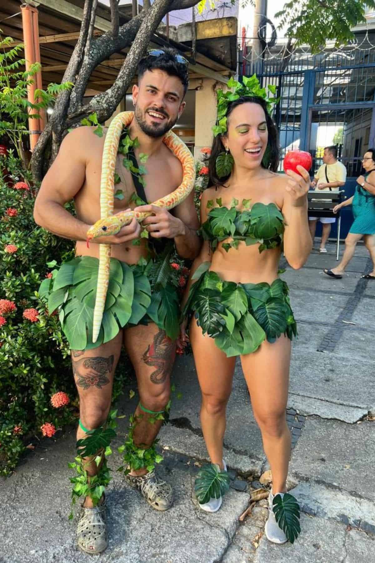 Two people dressed in Adam and Eve costumes made of leaves; the man holds a snake, and the woman holds a red apple. They are standing outside among plants and buildings.