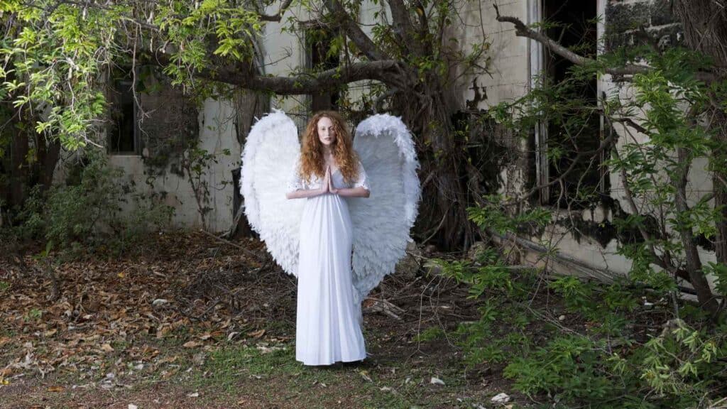 A person in a long white dress with large white angel wings stands outside near an overgrown, abandoned building.
