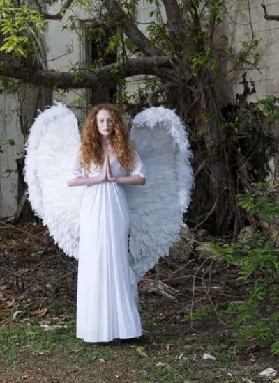 A person in a long white dress with large white angel wings stands outside near an overgrown, abandoned building.