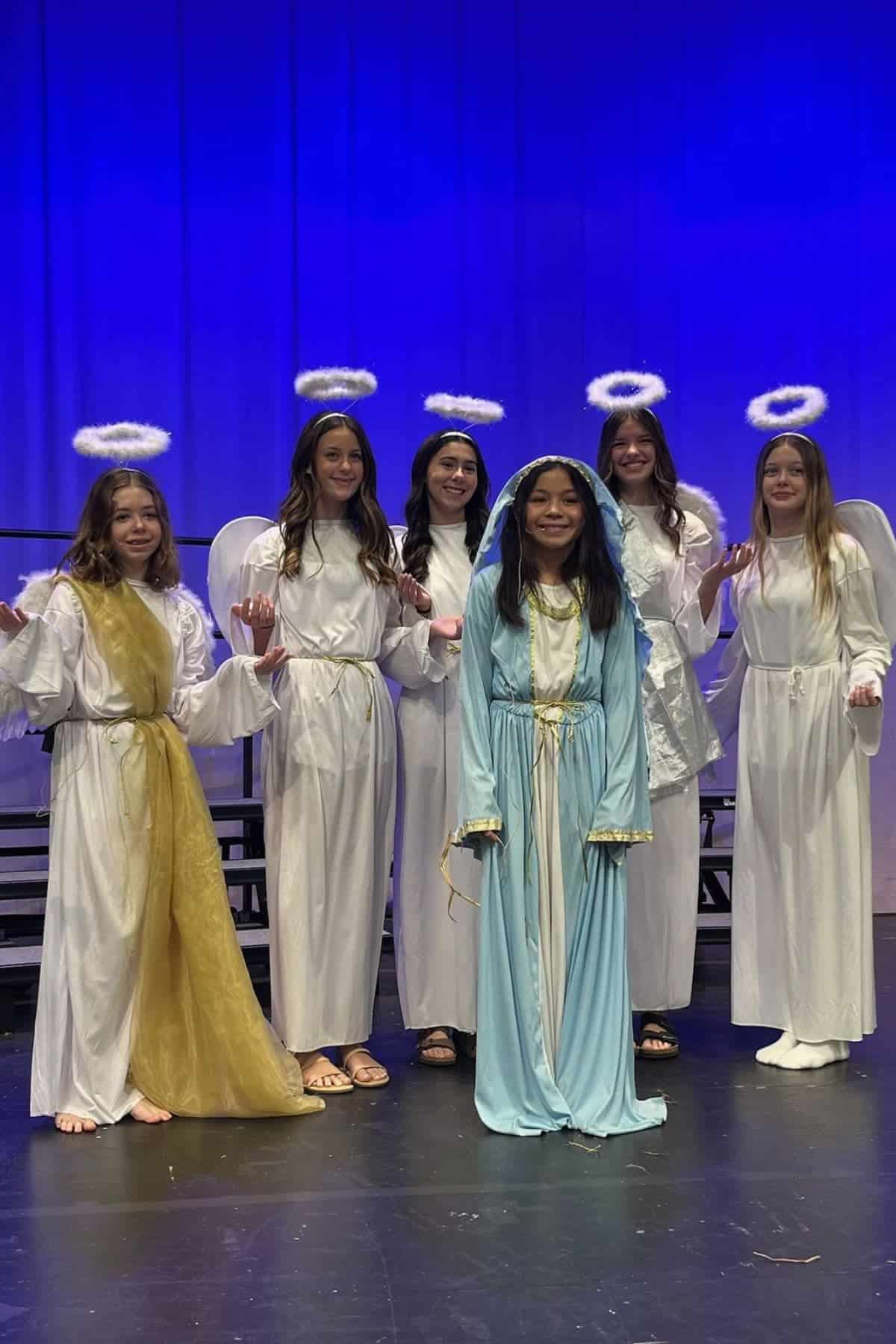 Six girls in costume stand on stage; five wear white robes and halos as angels, one in front wears a light blue dress resembling Mary. Blue and purple stage lighting in the background.