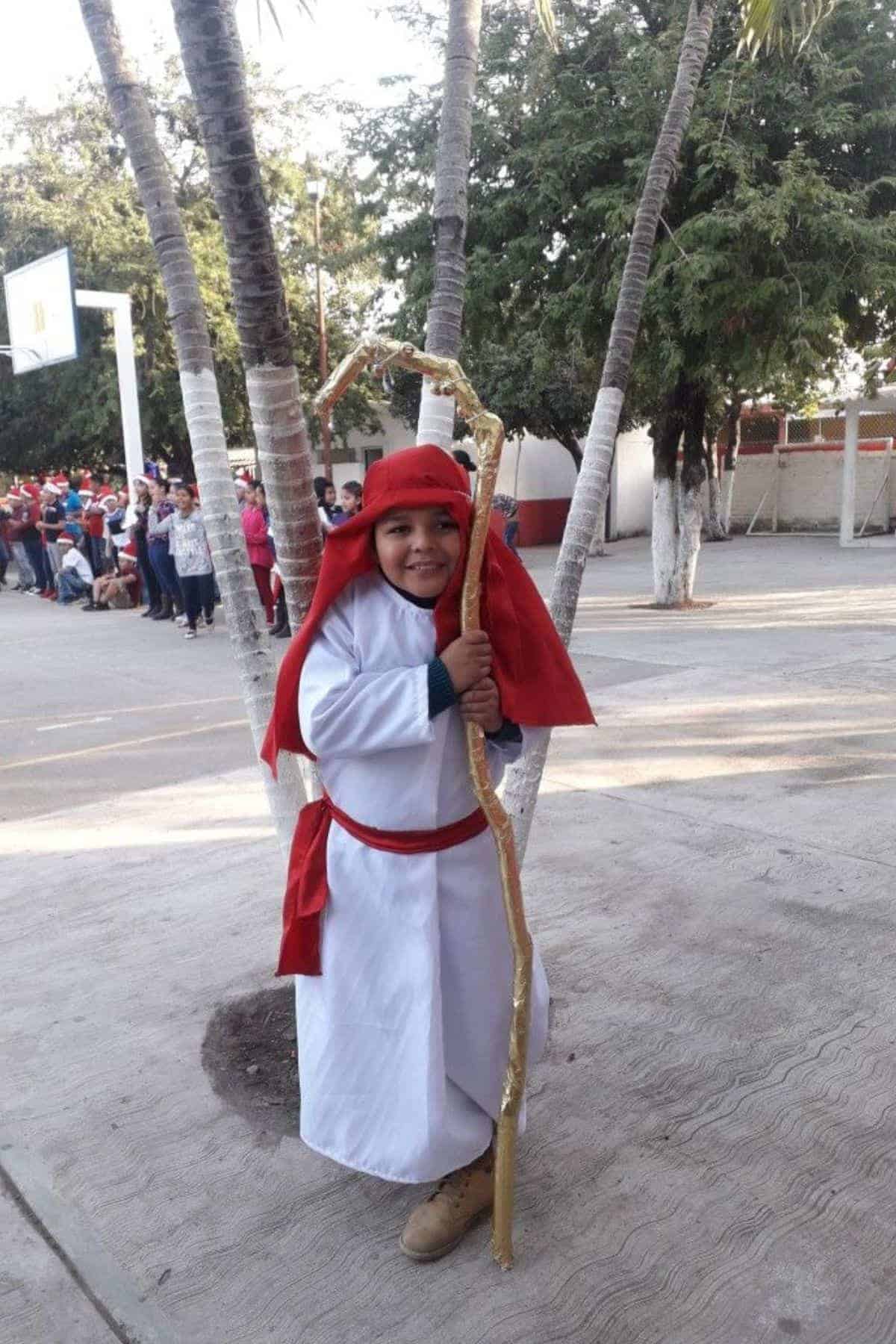 A child in a white robe and red head covering holds a staff, standing outdoors near trees, with a crowd in the background.
