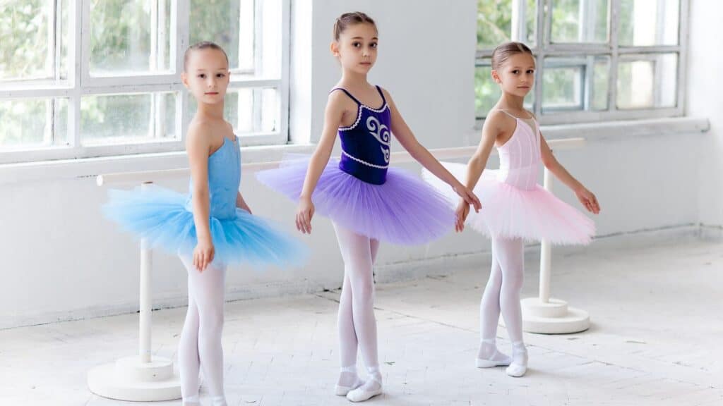 Three young ballet dancers in tutus and tights stand at a barre in a bright studio, posing in first position.