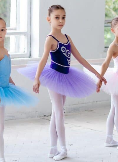 Three young ballet dancers in tutus and tights stand at a barre in a bright studio, posing in first position.