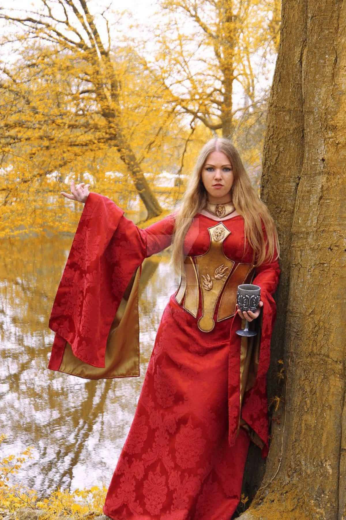 A woman in a red and gold medieval-style dress stands by a tree near water, holding a goblet and raising one hand. Autumn trees and reflections are visible in the background.