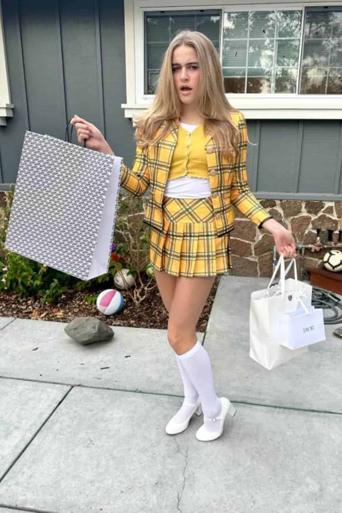 Young woman in a yellow plaid skirt suit stands outdoors holding shopping bags, wearing knee-high white socks and white shoes, with a surprised expression.