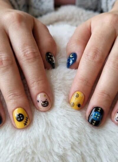 Two hands rest on a white fuzzy surface, displaying fingernails painted with colorful designs including cats, stars, and cosmic patterns in blue, yellow, and black.
