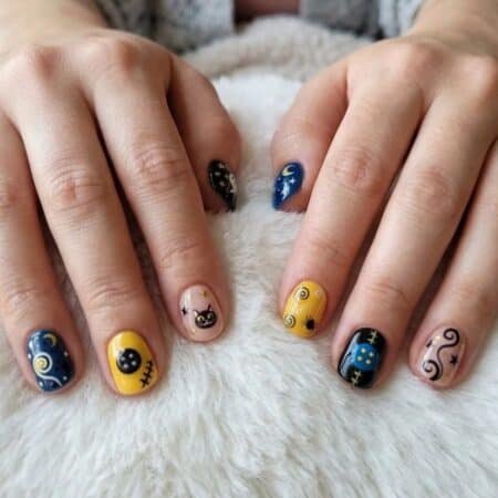Two hands rest on a white fuzzy surface, displaying fingernails painted with colorful designs including cats, stars, and cosmic patterns in blue, yellow, and black.
