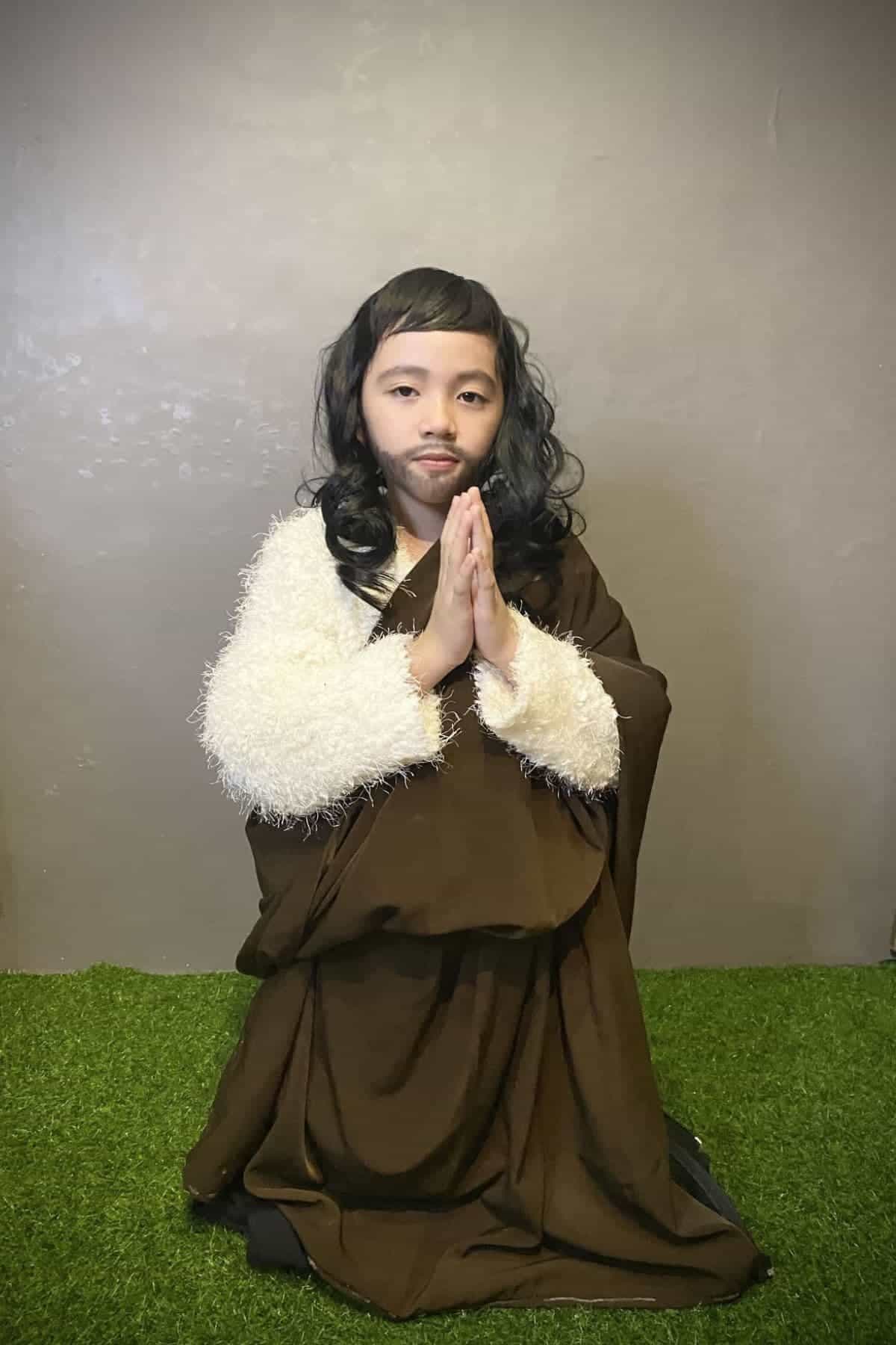 A child in a brown robe and white sleeve kneels on green carpet, hands pressed together in prayer, against a plain beige wall.
