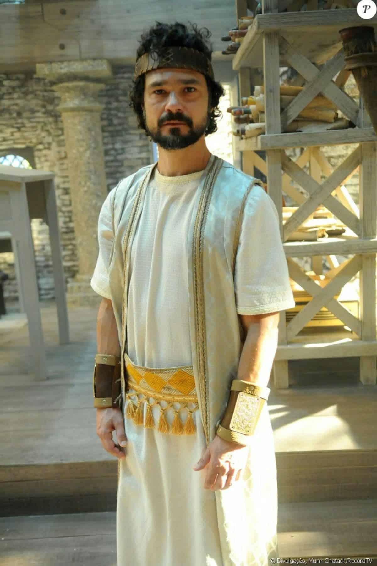 A man with a beard stands indoors wearing a light robe with gold accents, wrist cuffs, and a headband. Shelves and stone pillars are visible in the background.