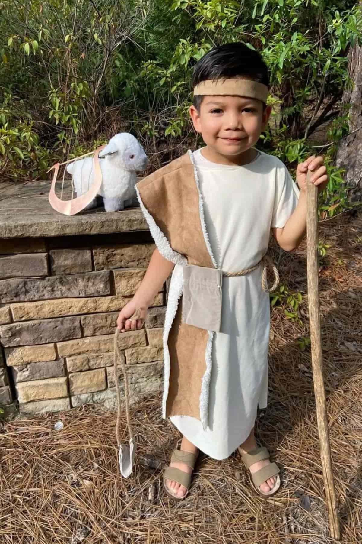 Young child in a simple costume holds a staff and sling, standing outdoors near a stone wall, with a plush sheep beside him.