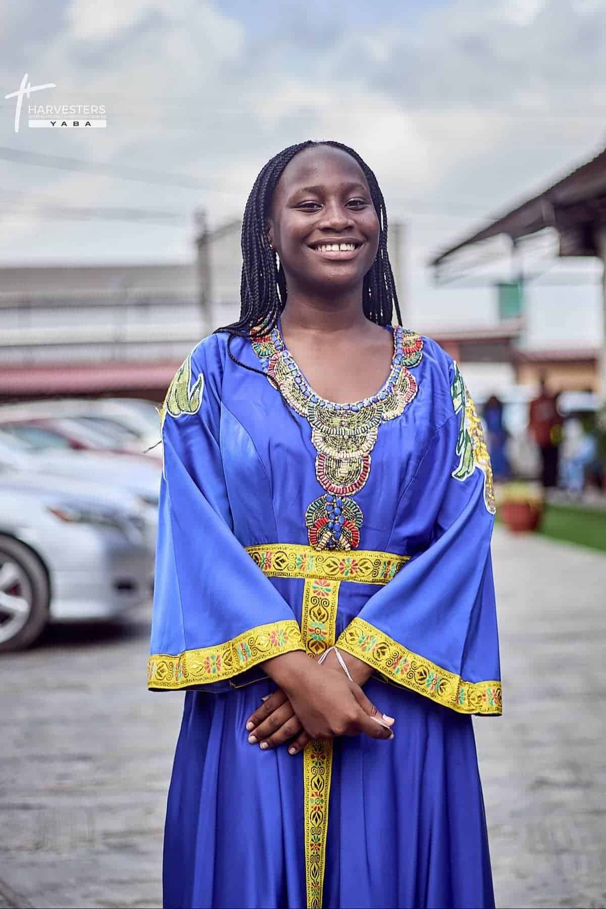 A woman in a blue and yellow traditional dress stands outdoors, smiling with hands clasped. Cars and buildings are visible in the background.