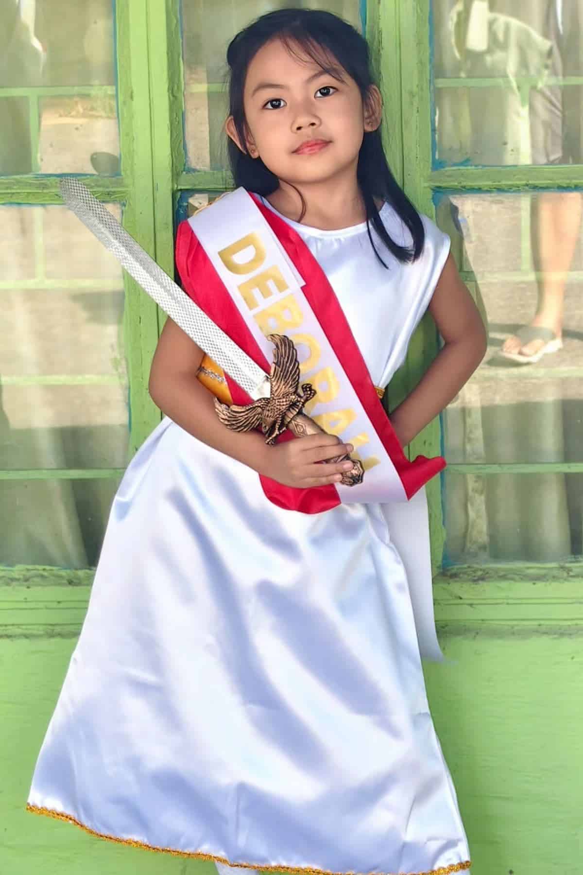 A young girl in a white dress with a “Deborah” sash and a toy sword poses in front of green window panels.