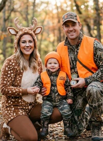 Three people pose in the woods; one is dressed in a deer costume, the others wear camouflage hunting gear and orange vests. Autumn leaves cover the ground and trees.