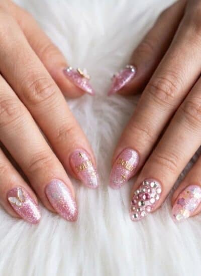 Two hands with long, almond-shaped nails featuring pink glitter polish and various decorative rhinestones and designs, resting on a white, fluffy surface.