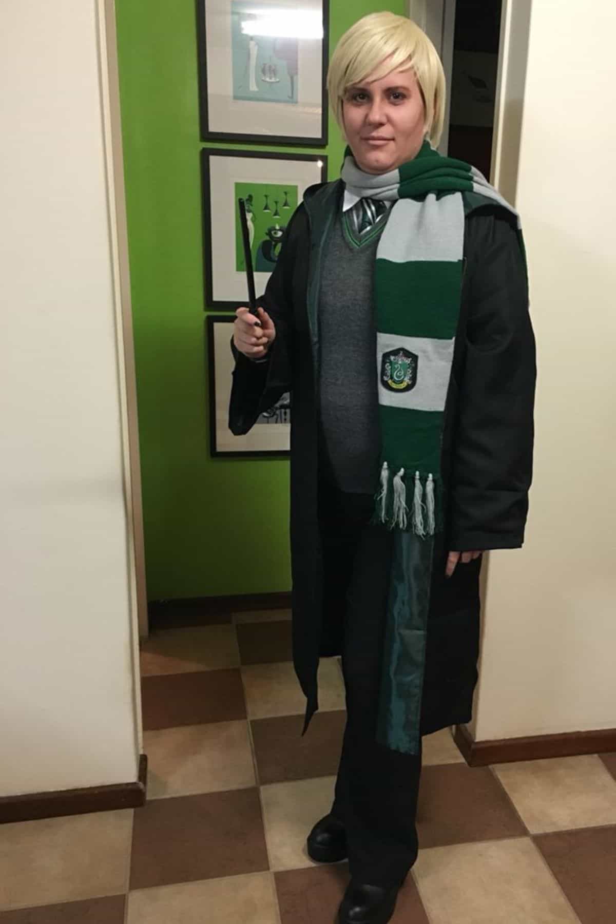 Person in a Slytherin costume with a green and silver scarf, black robe, and holding a wand, standing indoors on a checkered floor.