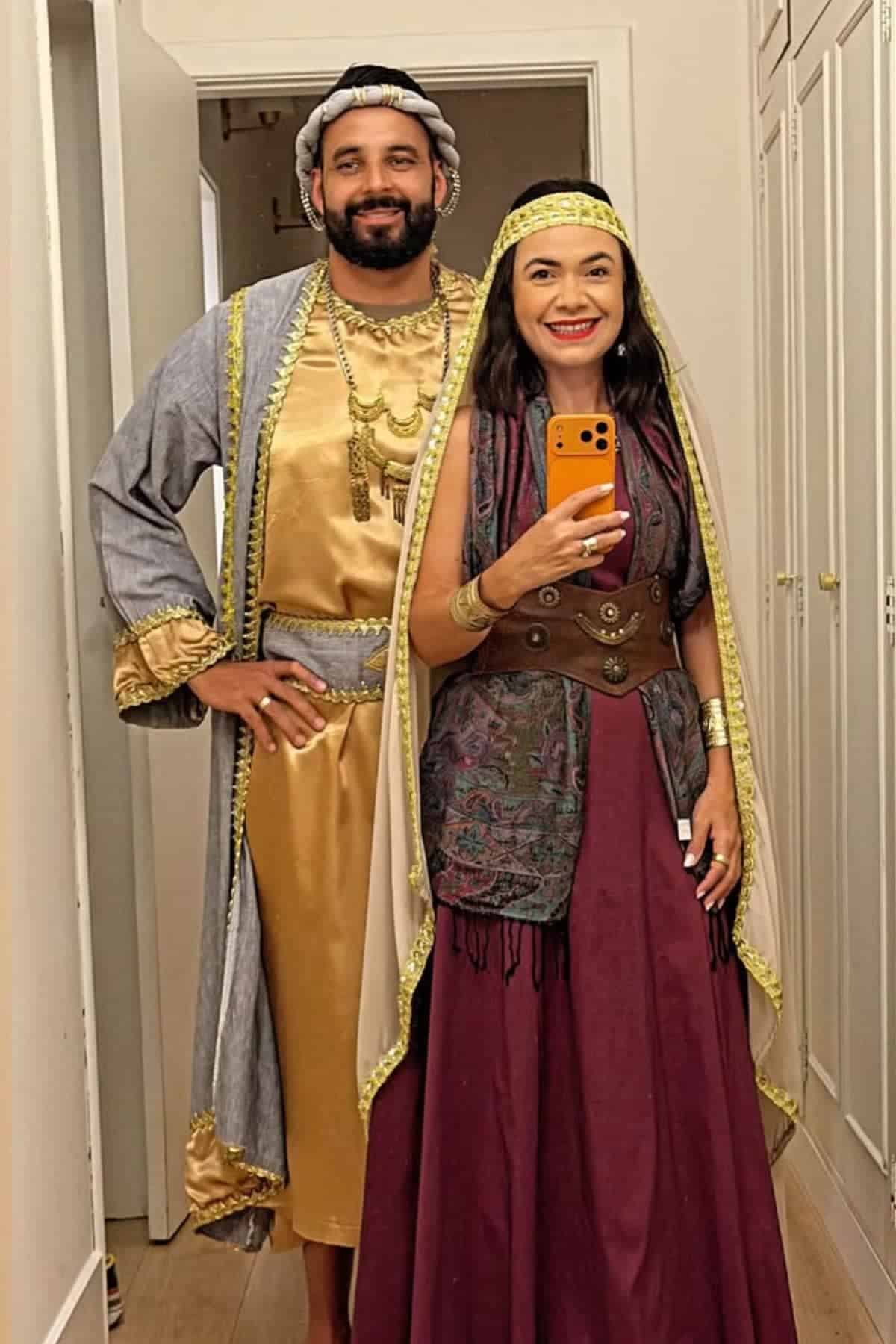 Two adults stand indoors wearing traditional or historical costumes, with one taking a mirror selfie on a smartphone.