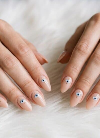 A pair of hands with light pink almond-shaped nails decorated with small blue and white evil eye designs, resting on a white fuzzy surface.
