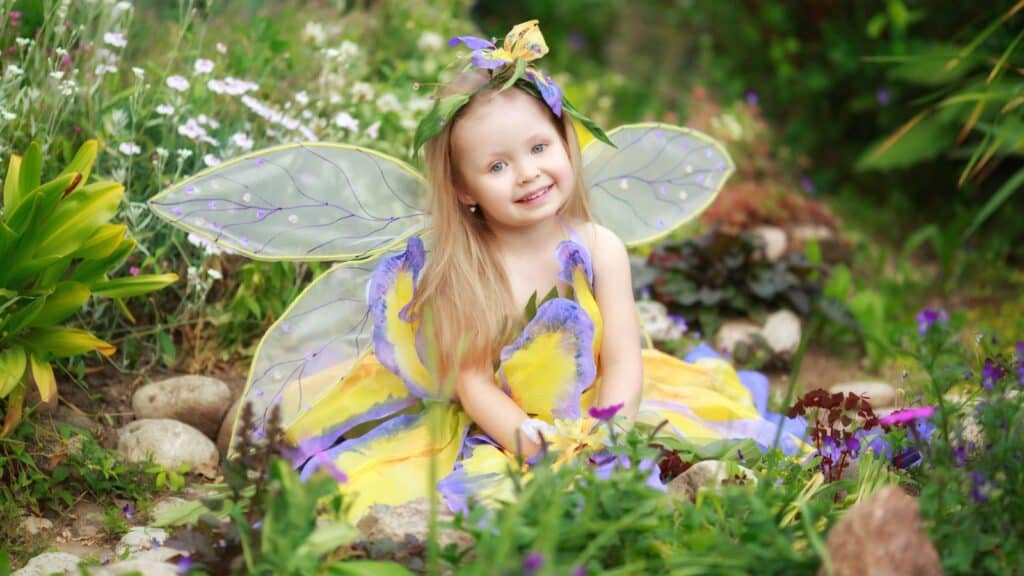 A young child dressed as a fairy with yellow and purple wings and a matching dress sits among flowers and greenery, smiling at the camera.