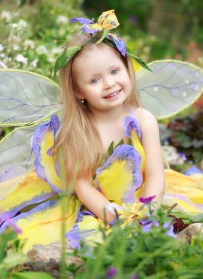 A young child dressed as a fairy with yellow and purple wings and a matching dress sits among flowers and greenery, smiling at the camera.