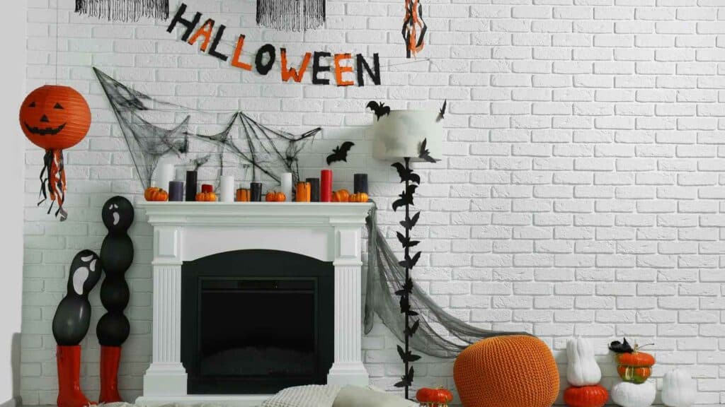 White brick wall with Halloween decorations including a banner, cobwebs, bats, pumpkins, candles, black and orange balloons, and a jack-o’-lantern lantern around a white fireplace.