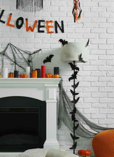 White brick wall with Halloween decorations including a banner, cobwebs, bats, pumpkins, candles, black and orange balloons, and a jack-o’-lantern lantern around a white fireplace.