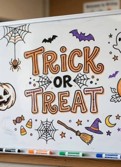 A whiteboard with “Trick or Treat” written in orange and black, decorated with Halloween drawings like pumpkins, spiders, bats, a ghost, and stars. Four markers are on the tray below.