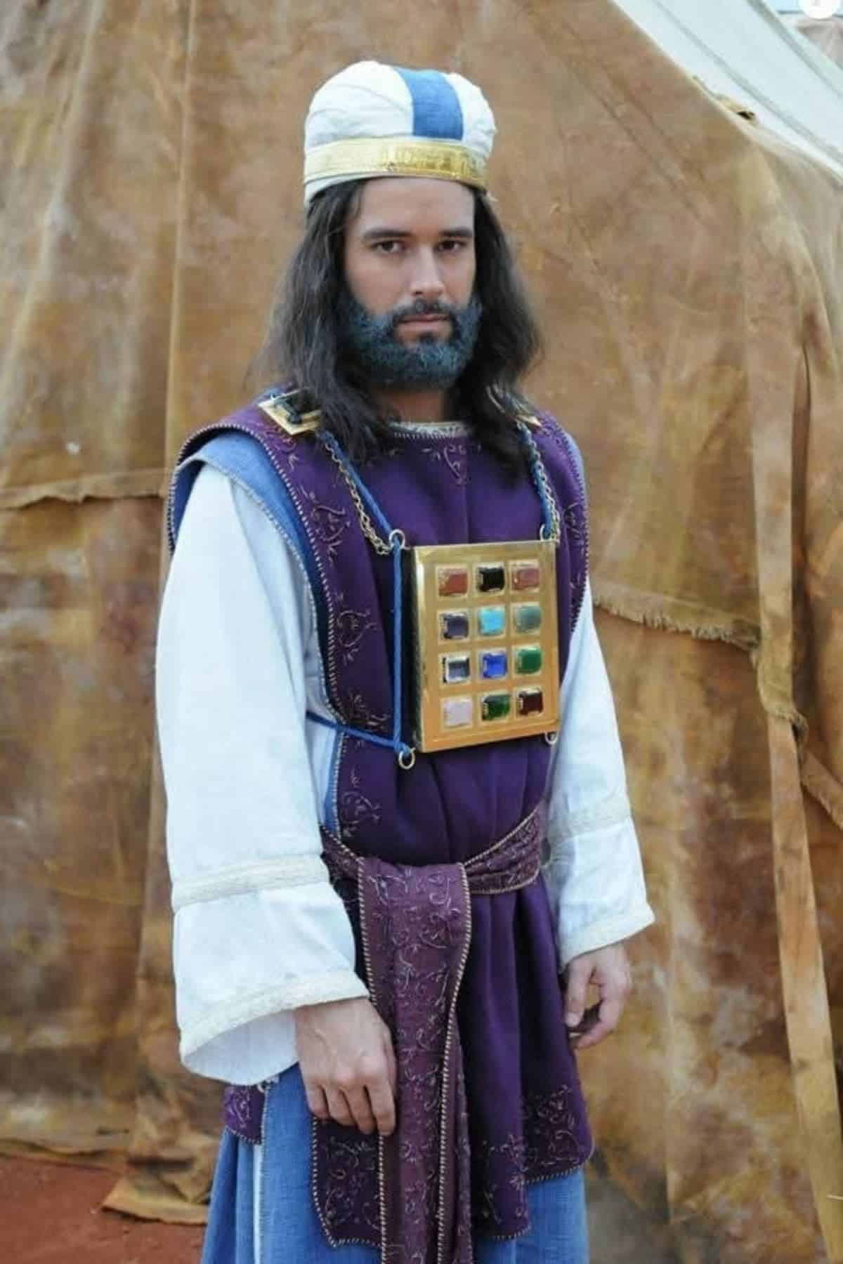 Man in biblical-era costume with a white and blue headdress, purple robe, and a breastplate featuring twelve colored stones, standing in front of a brown tent backdrop.
