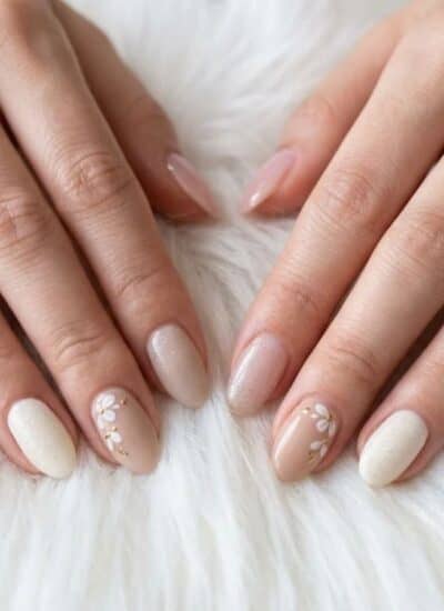 Two hands with manicured nails in neutral and pale pink shades, some featuring small white floral designs, resting on a white fluffy surface.