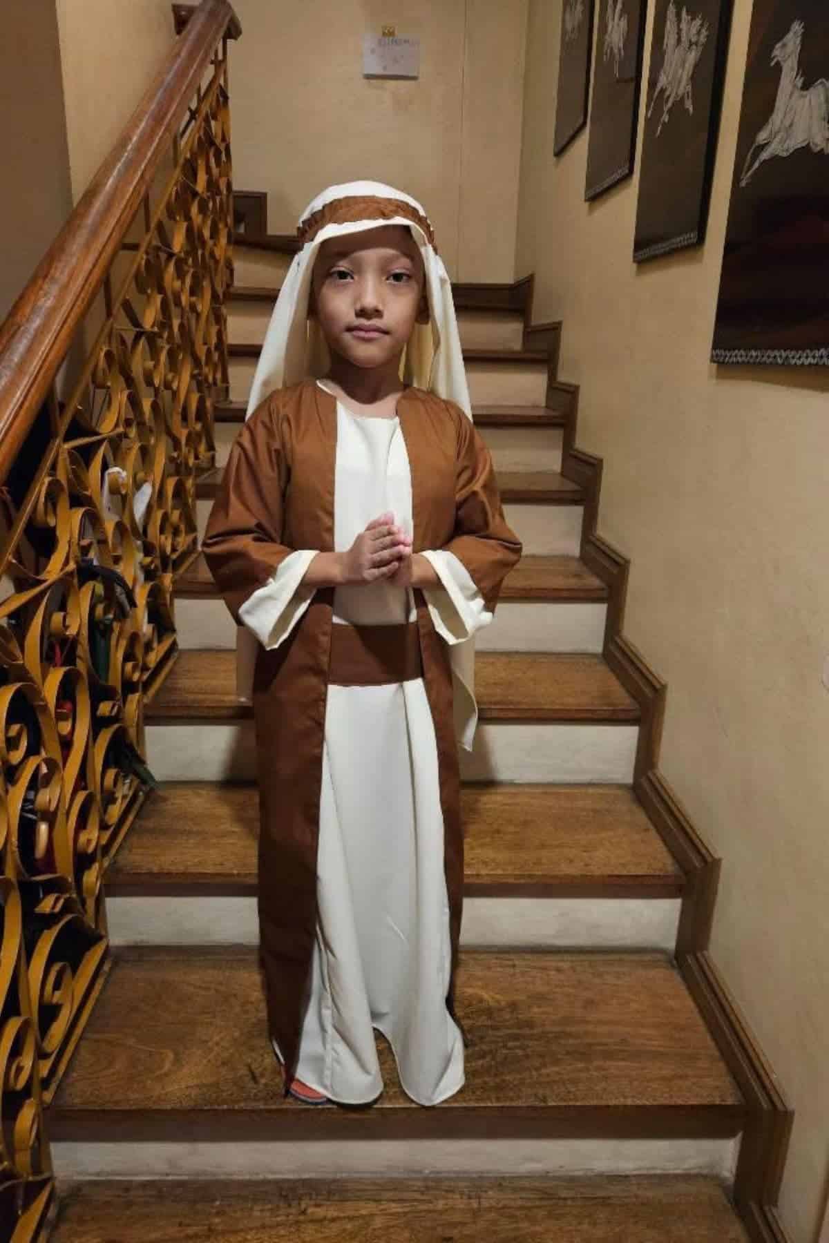 A child in a white and brown robe with a head covering stands on a wooden staircase, hands pressed together, facing forward.