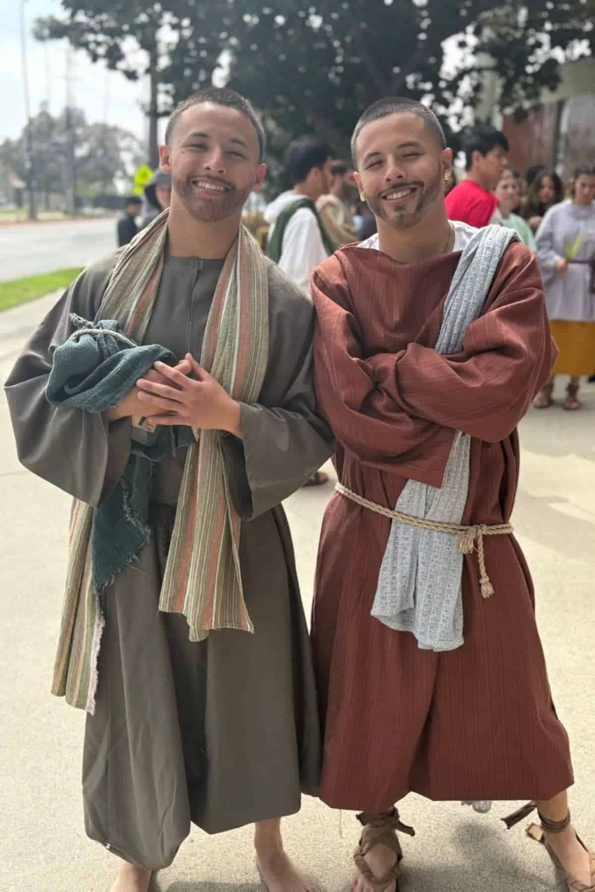 Two people wearing robes and sashes smile outdoors with a group of similarly dressed people in the background on a sunny day.