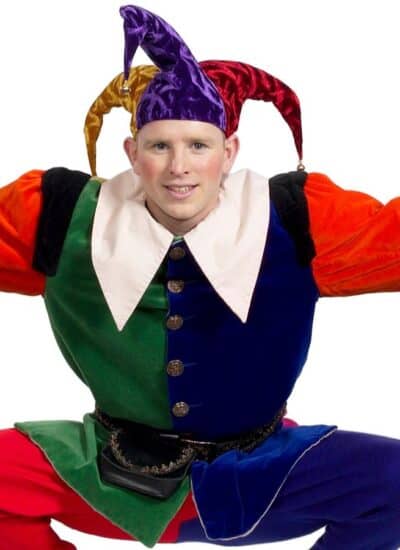 Person wearing a colorful jester costume with pointed hat, white collar, and mismatched colors, posing with arms bent outward and crouched legs.