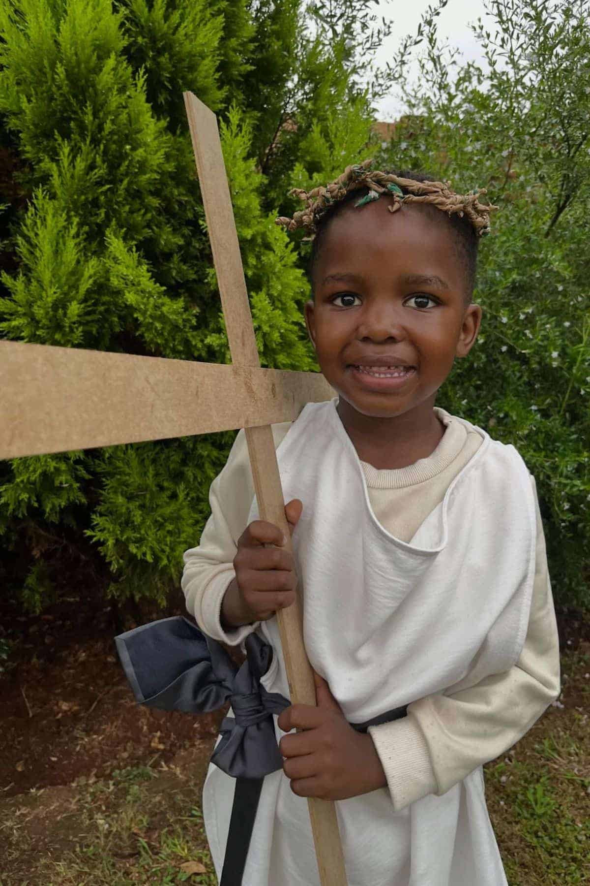 Smiling child dressed in a white robe and crown of thorns holds a wooden cross outside in front of green bushes.