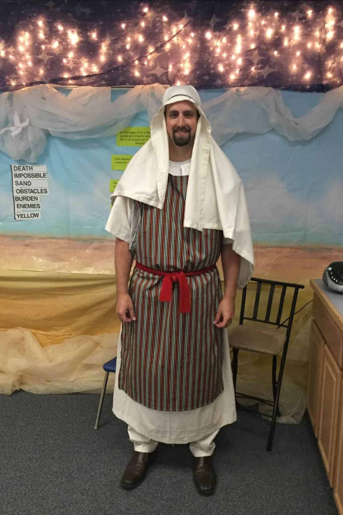 A man in a striped robe, white head covering, and red sash stands indoors under string lights, next to a sign with text and a backdrop of sand and sky decorations.