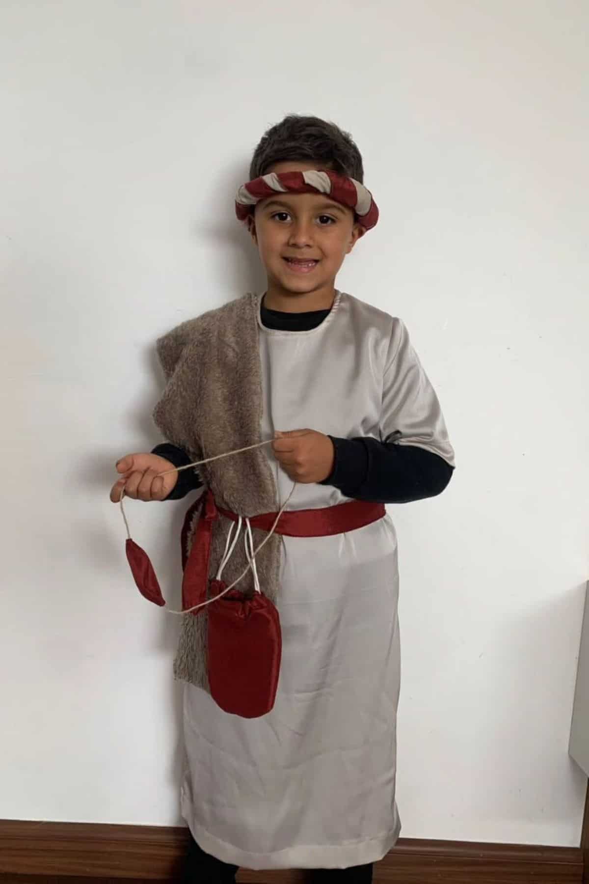 Child wearing a light-colored tunic, red belt, and headband, holding a homemade slingshot with red pouches, standing against a plain white wall.