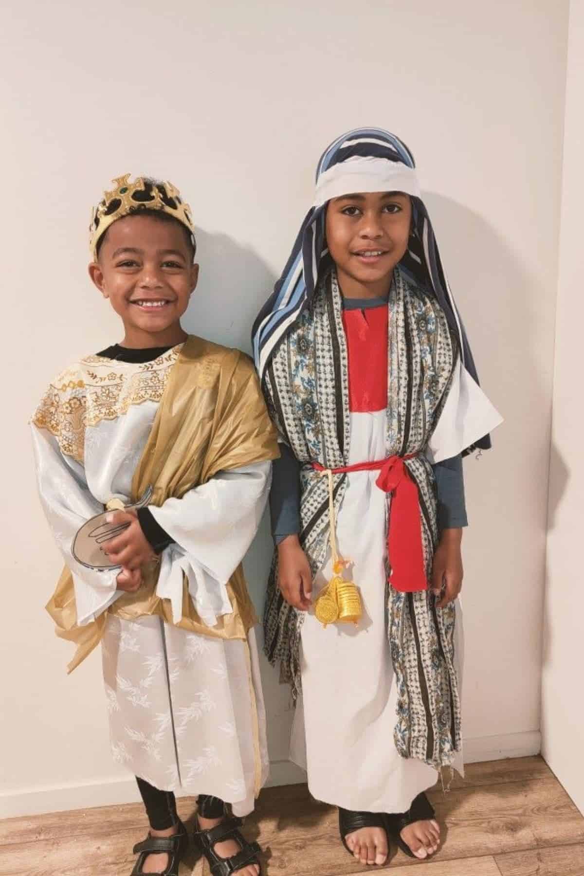 Two children stand side by side wearing gold and white costumes, including crowns and robes, against a plain white wall on a wooden floor.