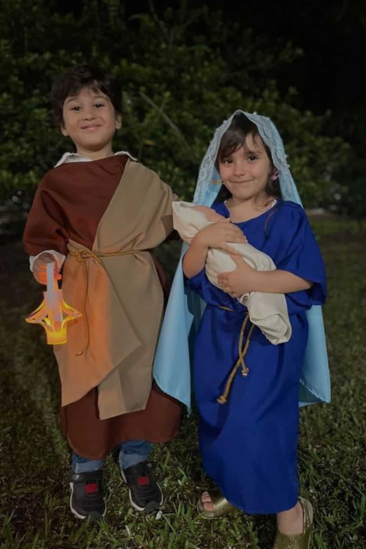 Two children dressed as biblical figures stand outside; one in brown holds a lantern, the other in blue holds a wrapped bundle, possibly representing Mary and Joseph in a nativity scene.
