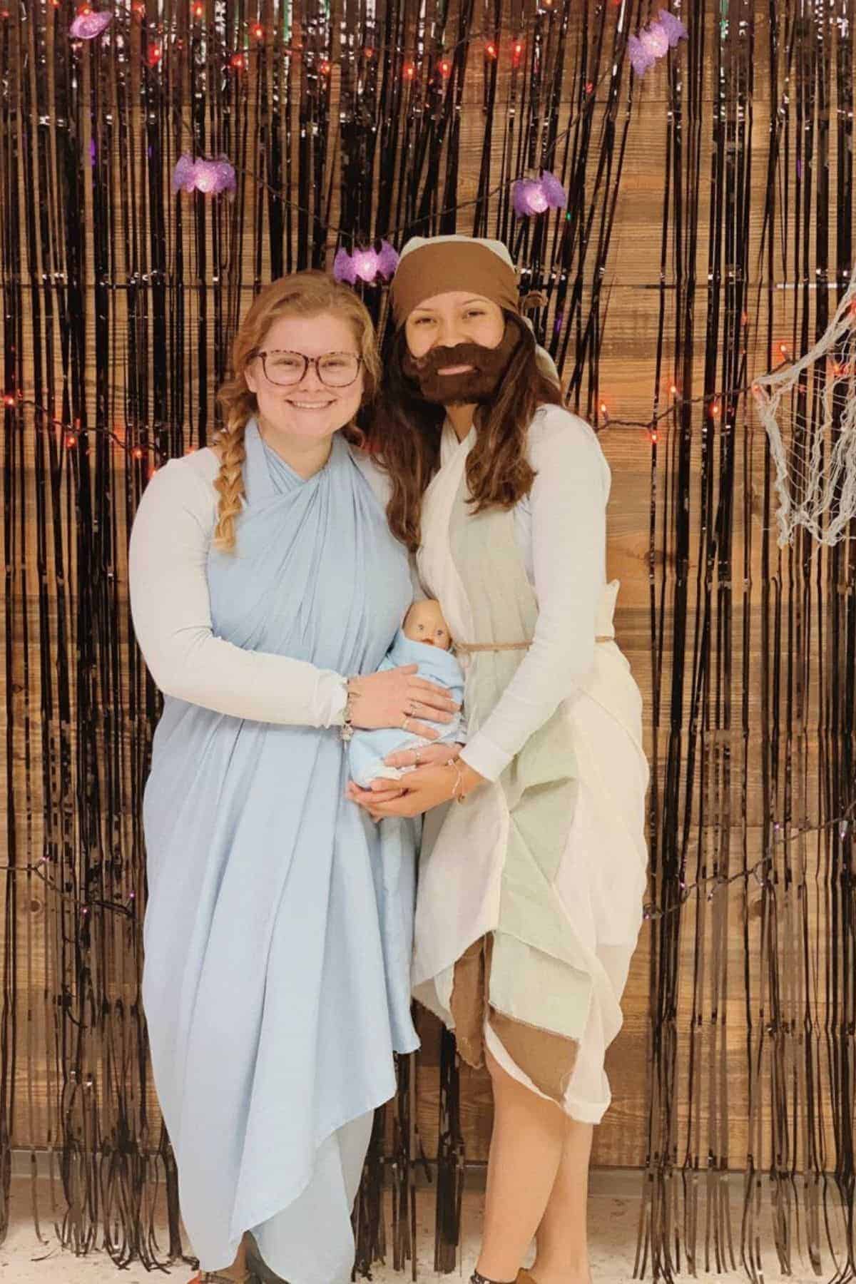 Two people dressed as Mary and Joseph hold a baby doll in a nativity-themed costume, standing in front of a backdrop with black streamers and purple lights.