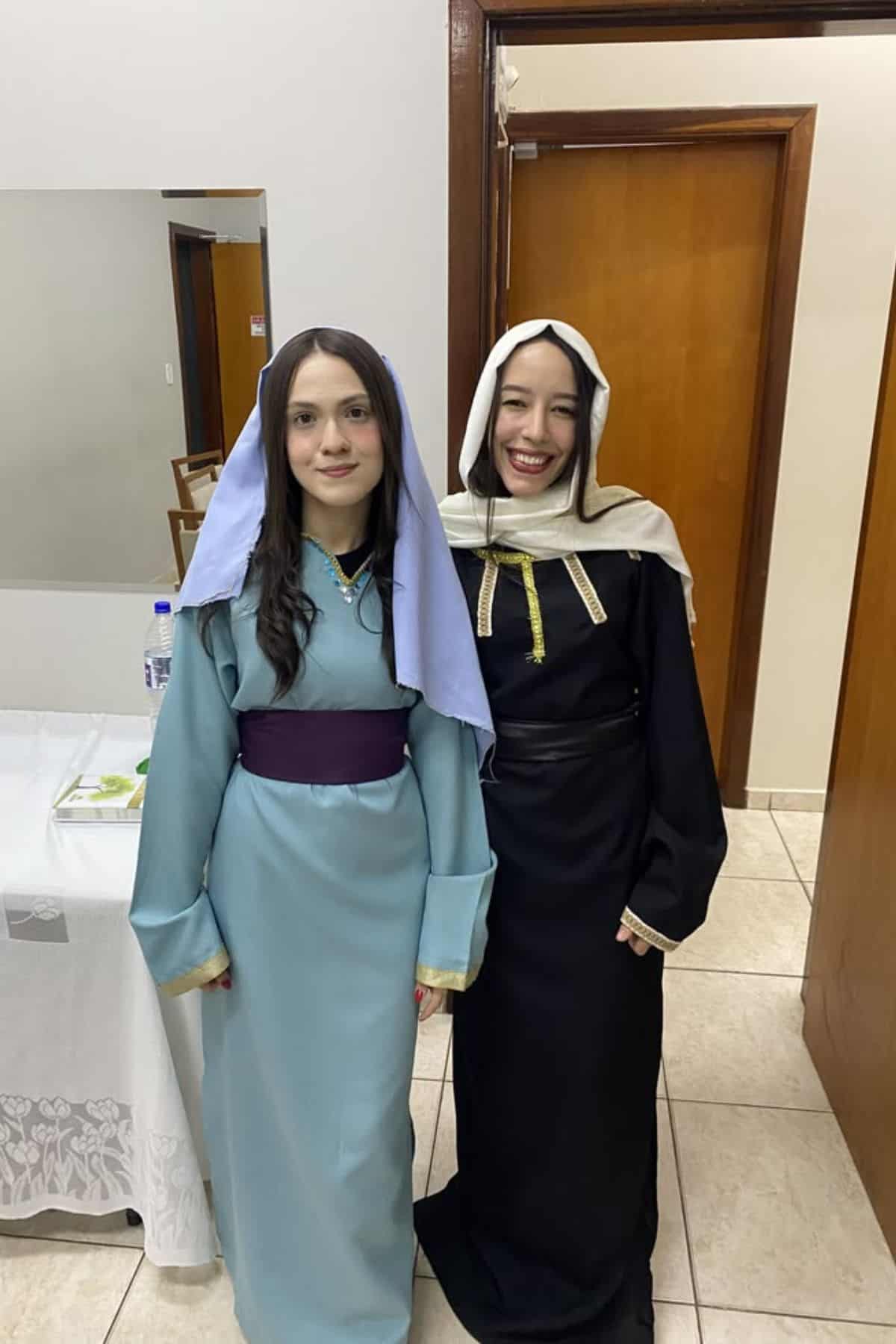 Two women indoors wear long robes and head coverings, standing side by side, smiling. One is in light blue with a purple sash, the other in black with gold trim.