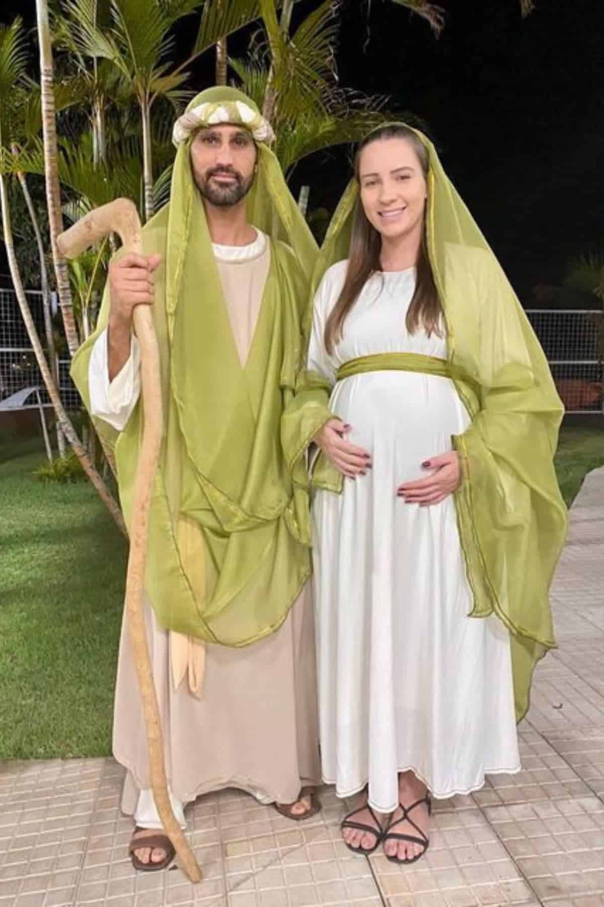 A man and a pregnant woman wear biblical-style costumes, standing outdoors at night, with palm trees and a fence in the background.