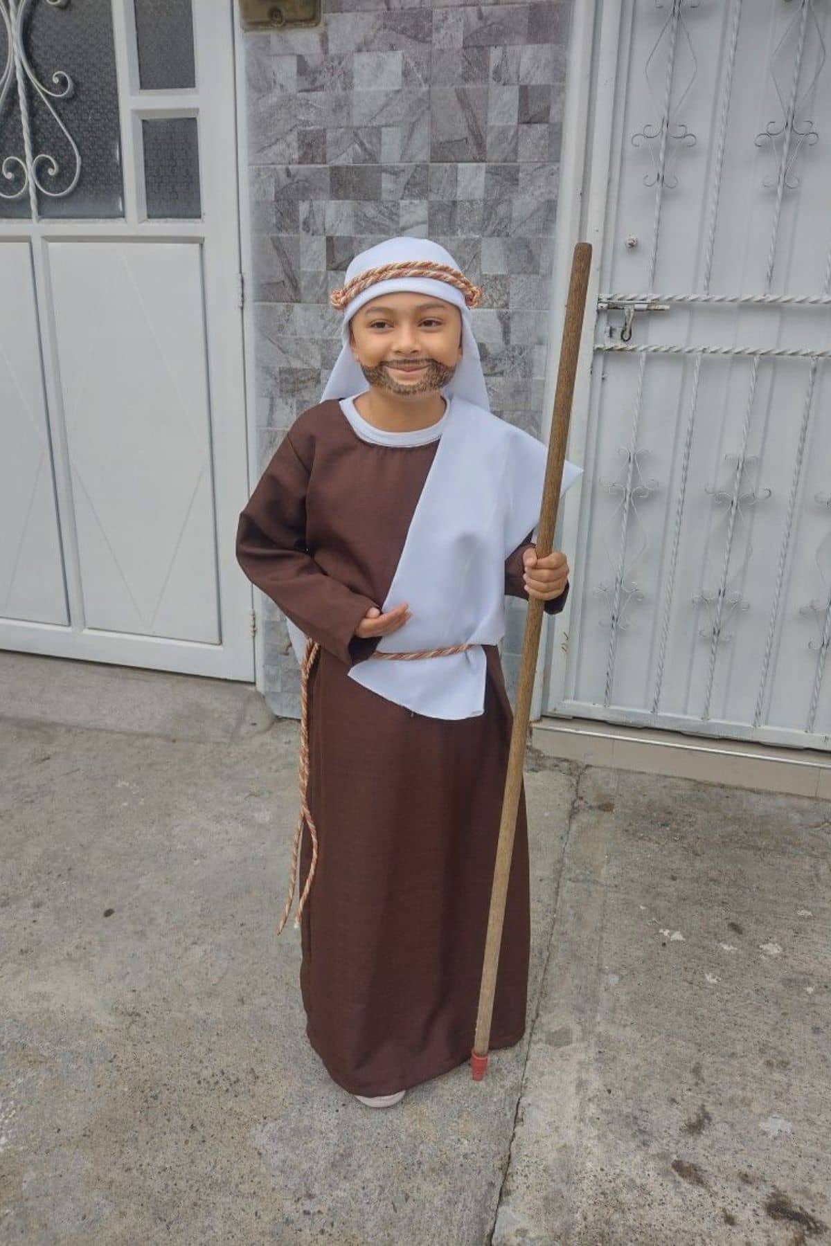 Child wearing a brown and white robe, head covering, and fake beard, holding a long stick, standing on a concrete surface in front of two doors.