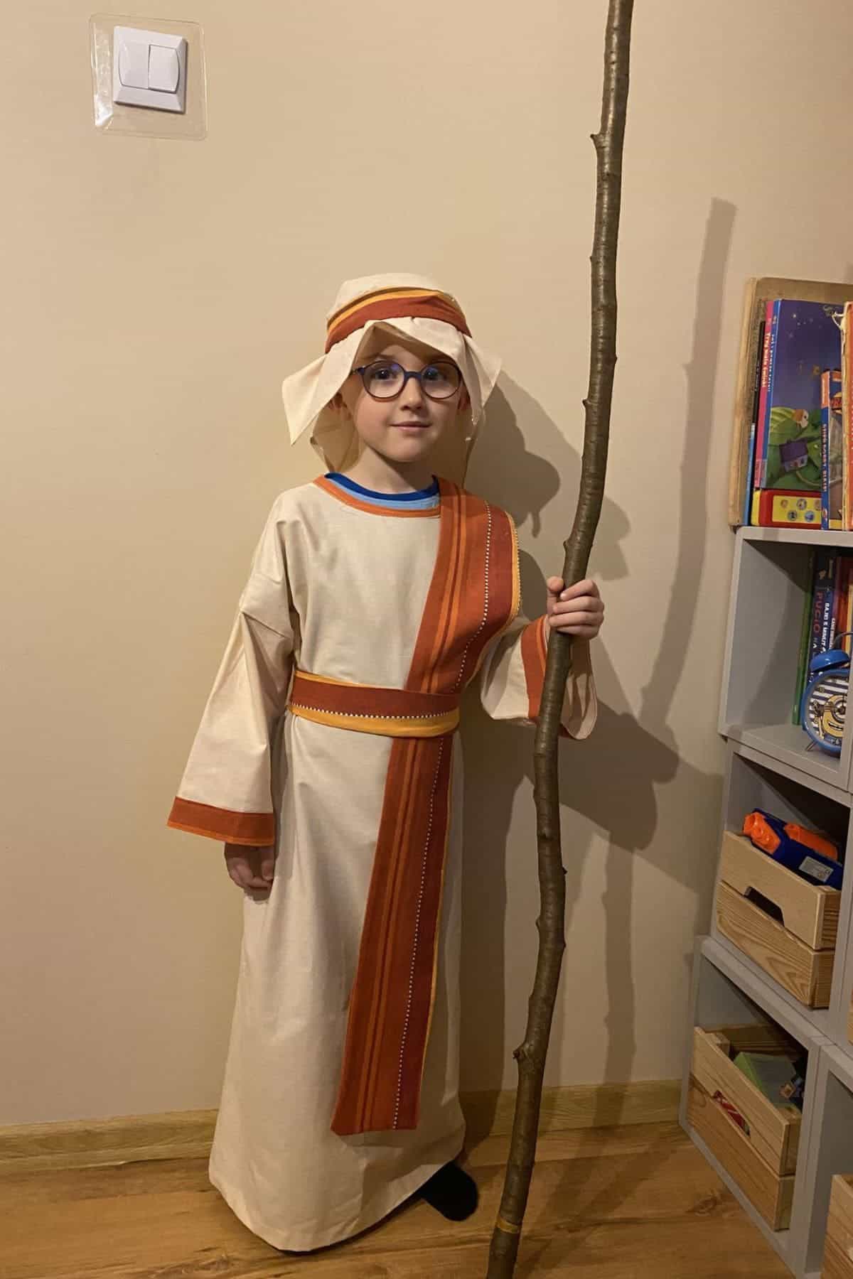 Child wearing a beige robe with an orange sash and head covering, holding a tall stick, standing indoors next to a shelf with books and toys.