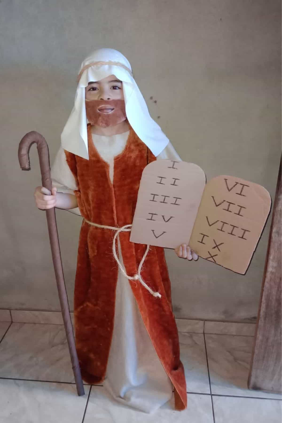Child in biblical costume holding a staff and two cardboard tablets with Roman numerals, standing indoors against a plain wall.