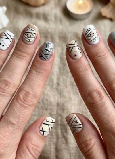 Two hands with Halloween-themed nail art, including mummy, ghost, and skeleton designs, are shown. Mini pumpkin, candle, leaves, and a small ghost figure are in the background on a beige cloth.