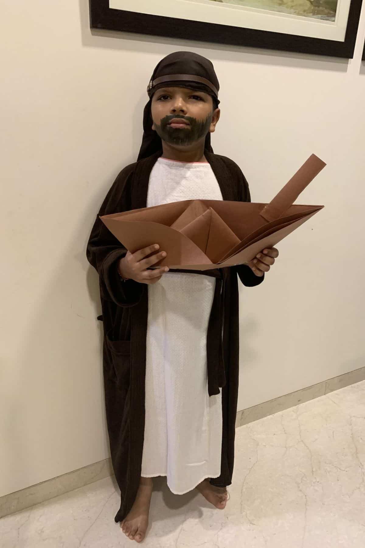 A child dressed in a robe, headscarf, and fake beard holds a large brown paper folded object, standing against an indoor wall.