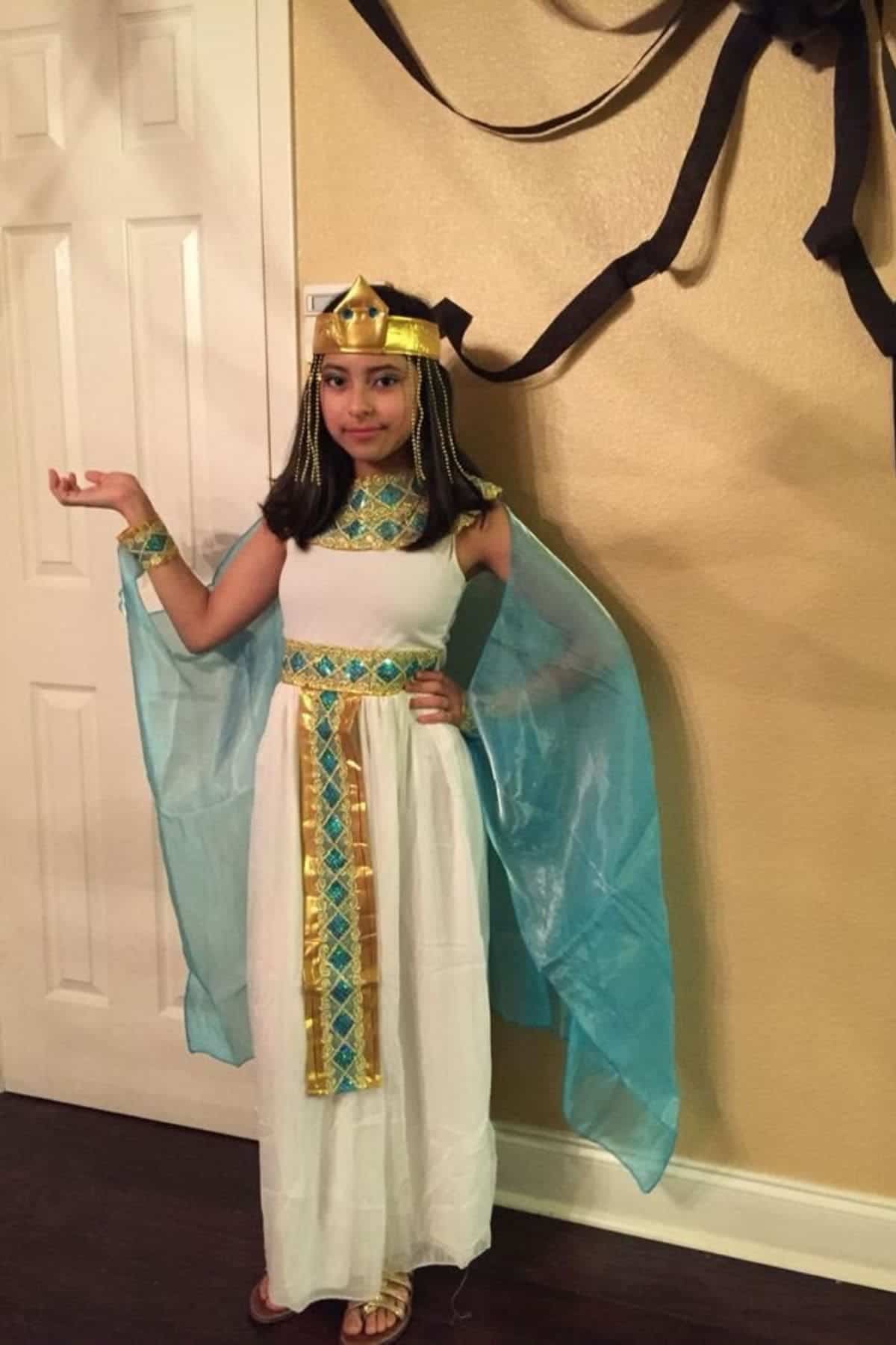 A young girl poses in an Egyptian-inspired costume with a gold headpiece, blue cape, and gold accents, standing in front of a tan wall with black decorations.