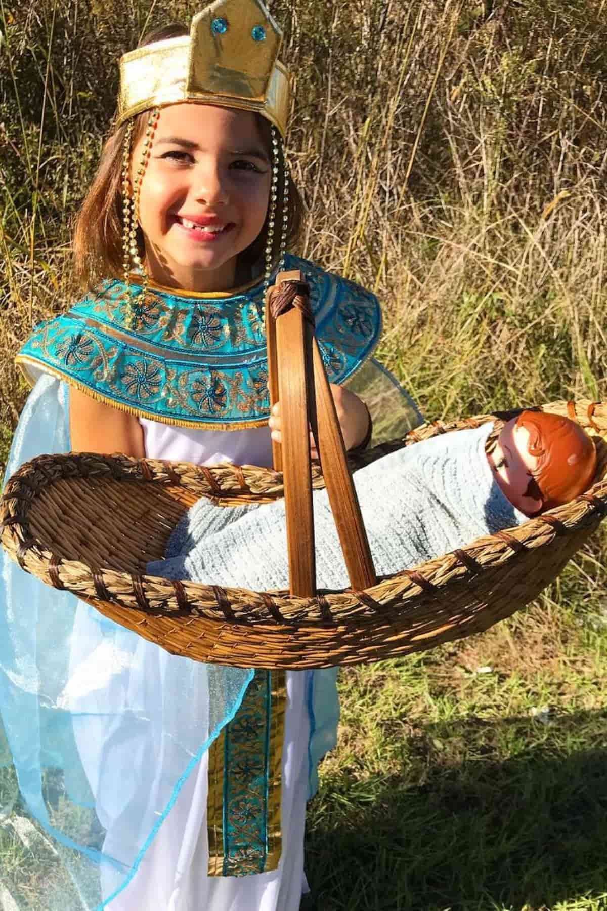 Smiling child in Egyptian costume holds a basket with a doll wrapped in a blanket, standing outdoors in a grassy area.