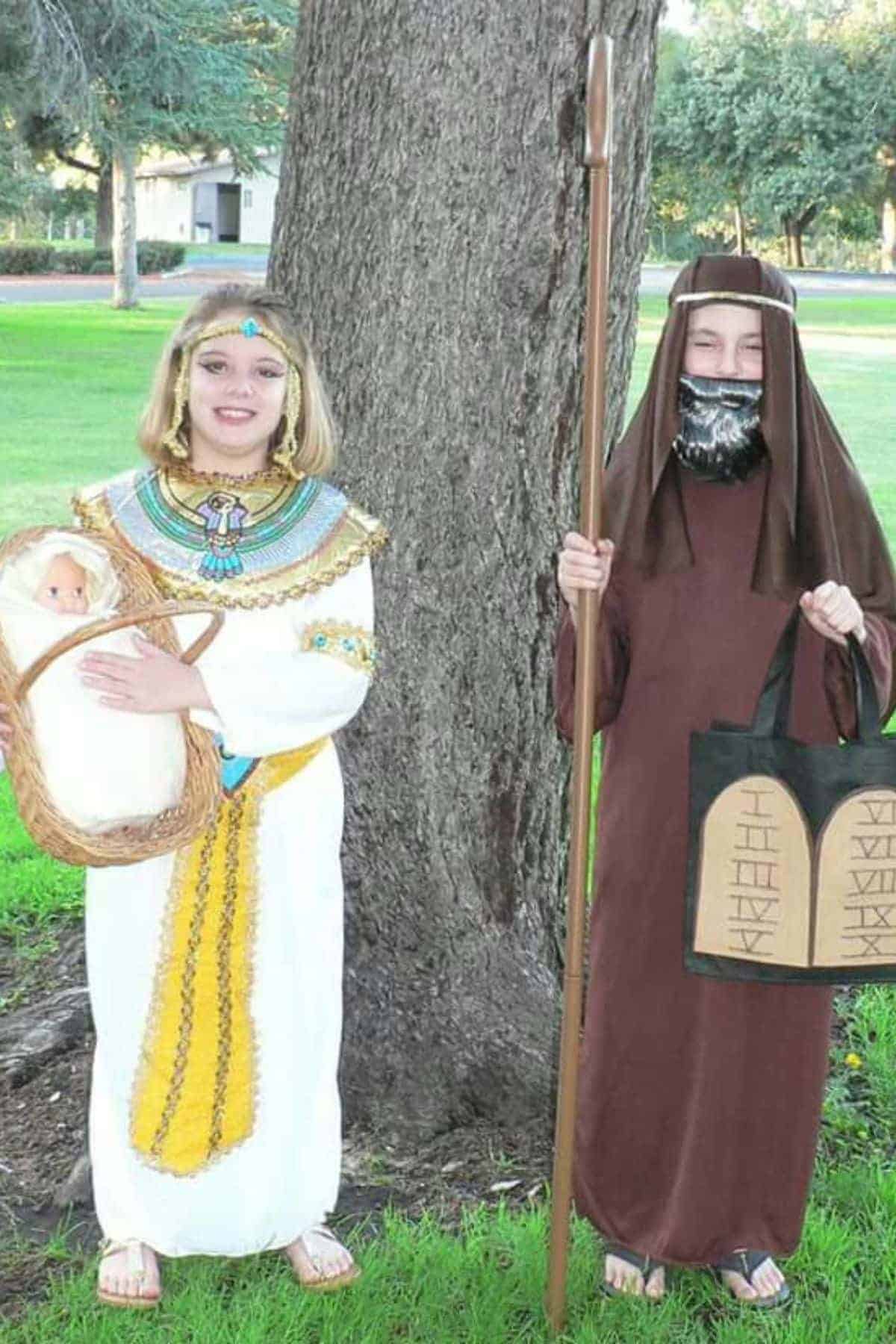 Two children in costumes stand outside; one is dressed as an Egyptian queen holding a baby doll, the other as a bearded figure in a robe holding a staff and two tablets.